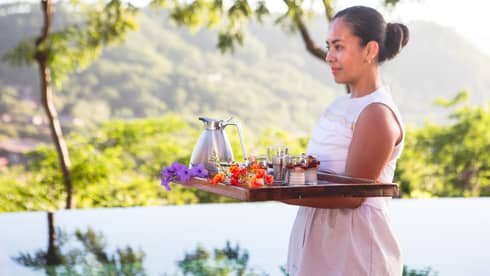Woman carries breakfast tray with tropical flowers past outdoor pool