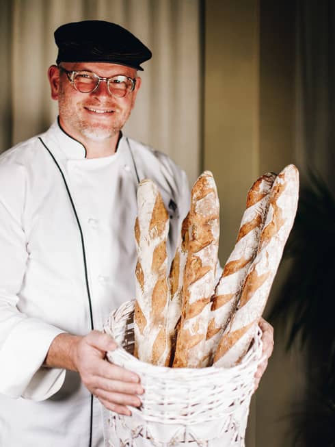 Baker Sven Mosteg, wearing a white chefs coat, glasses and a black beret, stands holding a basket of fresh-baked baguettes