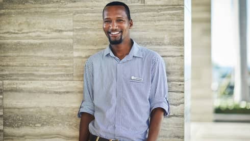 Smiling person in a checkered shirt standing against a stone wall indoors.