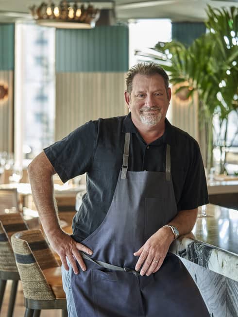 A chef in a black shirt and blue apron standing in a well lit restaurant.