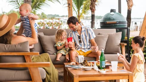 Family with two adults and three children ranging in age from infant to tween sit on grey outdoor sofas playing cards and enjoying food served on the coffee table in front of them