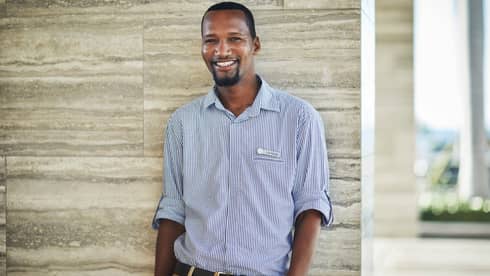 Smiling person in a checkered shirt standing against a stone wall indoors.