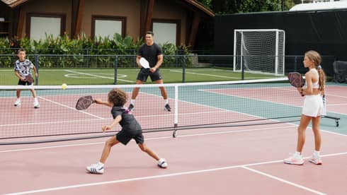 One adult and three children play pickleball on an outdoor court surrounded by trees