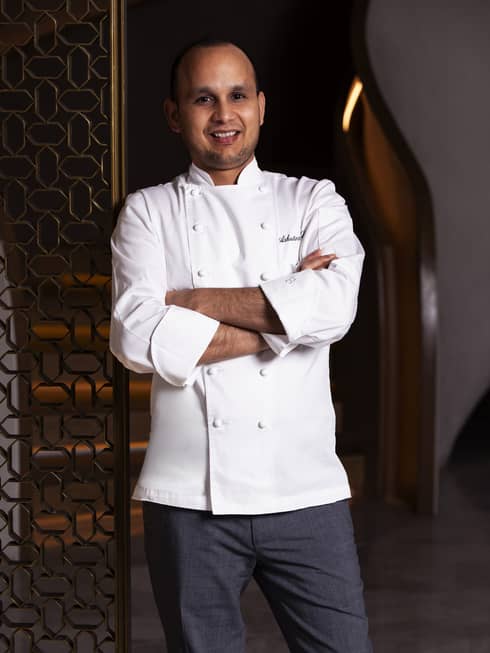 Chef with buzzed hair and light facial hair wearing a white chef jacket and dark-gray pants stands smiling with their arms crossed