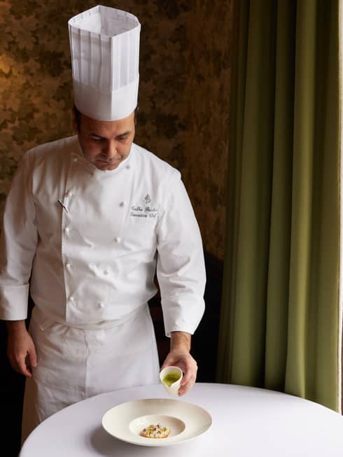 Chef Talha Barkin in white chef?s hat and uniform, standing beside a white table and pouring sauce on a food dish