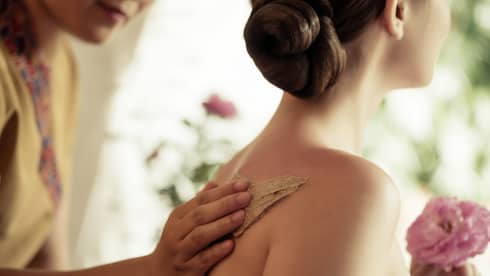 Spa staff rubs thick lotion on woman's bare shoulders as she holds a fresh flower