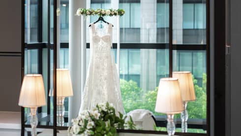 Reflection in a tall mirror of a wedding gown hanging across a window in a room with chandelier and white floral décor.