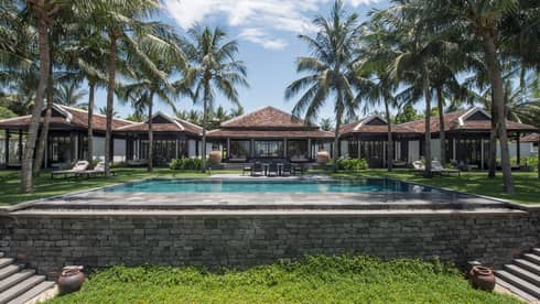 Luxurious villa with a large swimming pool, surrounded by palm trees and greenery, featuring modern architecture with tiled roofs and outdoor seating areas.