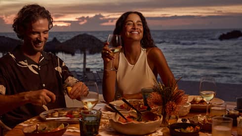 Man and woman laugh while dining at outdoor table filled with food and wine, next to beach and ocean