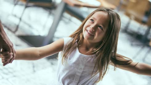 Close-up of girl with long hair and white dress, smiling up at adult who is holding her hand