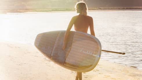 Silhouette of woman walking with large paddle board across beach at sunrise
