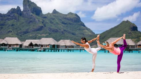 Two people practicing yoga on a tropical beach with overwater bungalows and mountains in the background