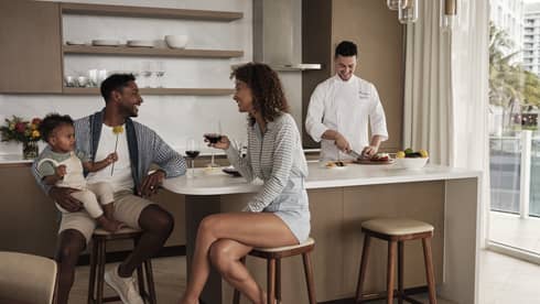 Parents sit at a kitchen island, toddler in dad's arms, laughing over glasses of red wine while a chef prepares a meal.
