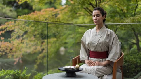 A woman sits on glass-enclosed balcony in lush setting with traditional Japanese robe on, meditating with an orin bell