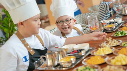 Two children wearing white chef's hats, white t-shirts and blue aprons sit at a table in front of various small plates of ingredients