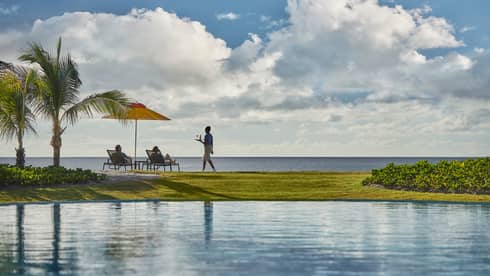 View across pool to green lawn, waitstaff carrying tray to couple in patio chairs overlooking ocean