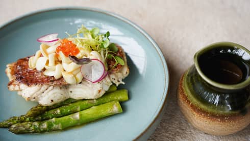 A pale blue plate holds a piece of white-fleshed fish placed next to asparagus spears and topped with garnishes. A green and tan glazed ceramic vessel sits next to the plate.