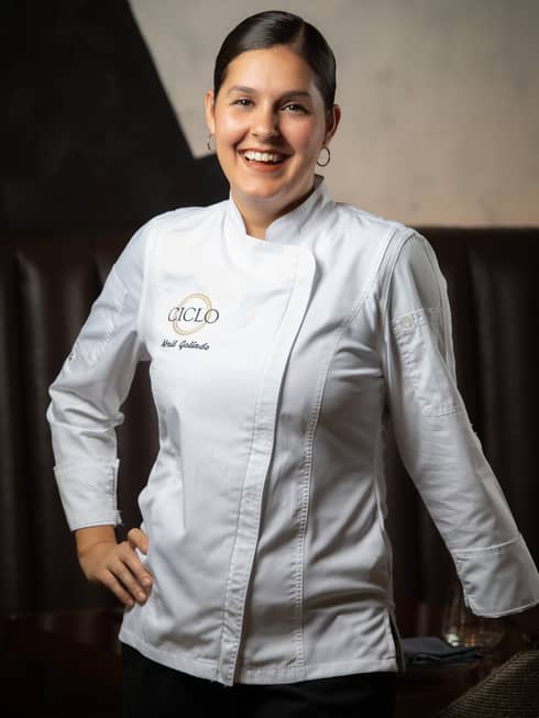 A portrait of female Chef Abril Galindo leaning against a table and smiling in her white chef coat