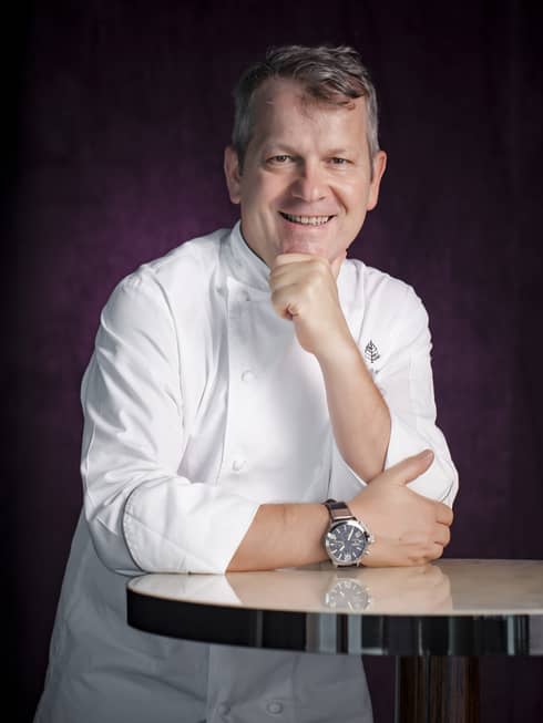 Smiling man wearing white chef's coat resting one arm on a round table and the other under his chin
