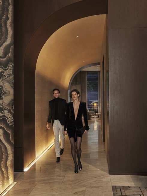 Elegantly dressed couple walking through a dimly lit, luxury hotel hallway with arched ceiling and marble accents