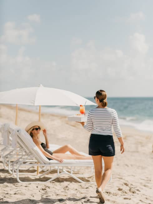 Woman carries tray with fruity cocktail to guest in lounge chair on sand beach