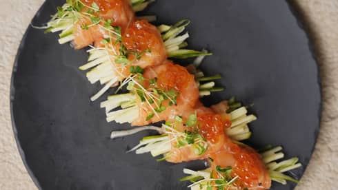 Five salmon rolls, topped with orange caviar and microgreens, in a row on black dish