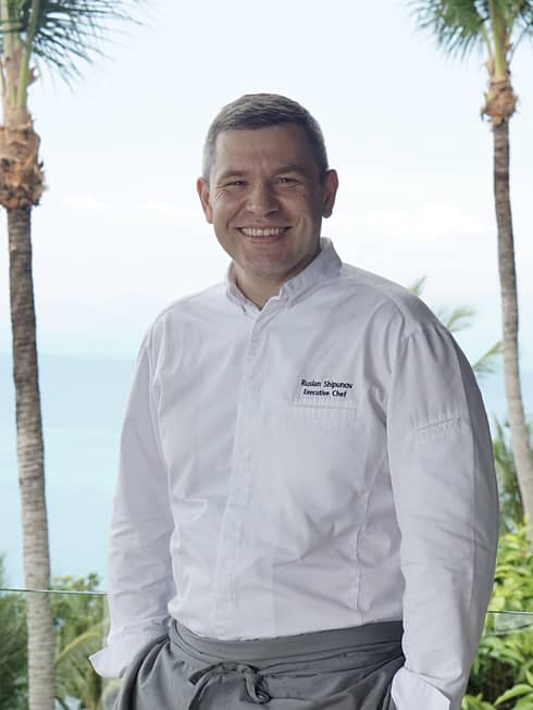 A smiling Executive Chef Ruslan Shipunov stands for an outdoor portrait flanked by two palm trees