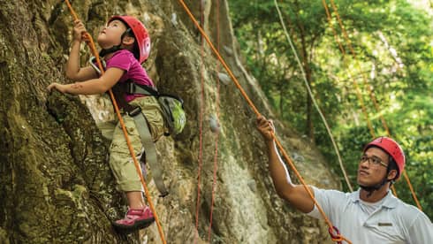 Man holds rope, helps young girl wearing helmet climb rock face