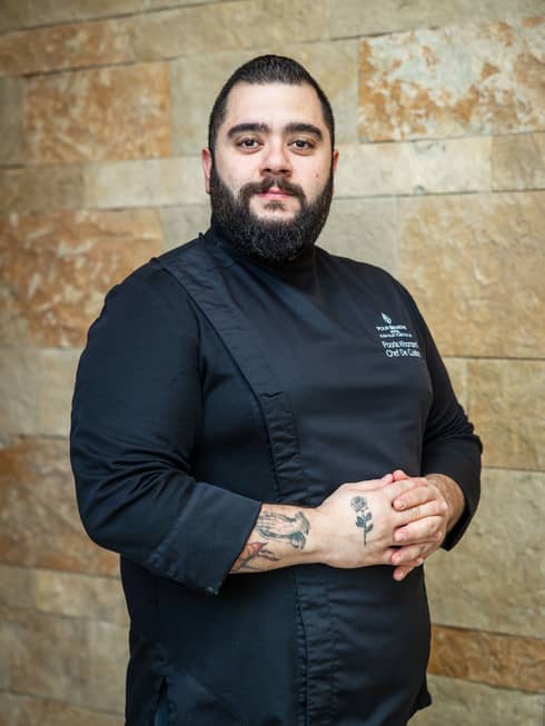 Person with short dark hair, trimmed dark beard and tattoos on his arms stands wearing an all-black chef's uniform in front of a stone wall