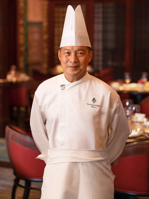 Person in white chef's coat and hat stands with hands behind back