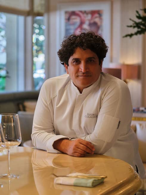 Chef with short, dark curly hair and wearing a white long-sleeve chef's jacket sits at a restaurant counter