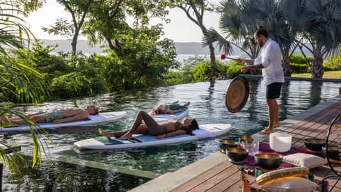 Three people float on stand-up paddleboards in a private pool while someone plays a gong on the deck