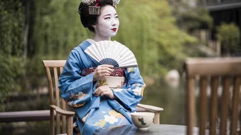 Geiko, a traditional Japanese entertainer, dressed in kimono and holding fan, sitting with cup of tea in garden
