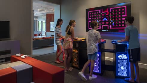 Four smiling teenagers stand at multi-coloured video-game pedestals facing a large TV screen in a dimly-lit games room.