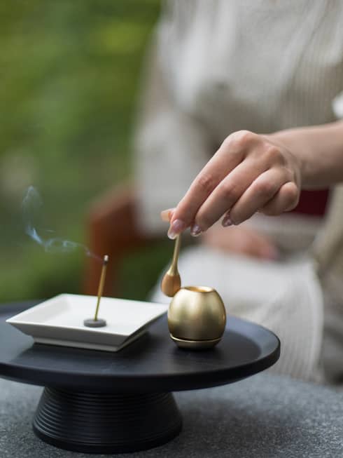 Close-up of a hand holding a small brass spoon over an orin bell and incense burner, with lit incense stick emitting smoke
