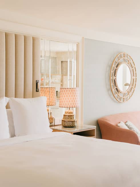 Elegant hotel room with a plush white bed, mirrored headboard, stylish lamps on nightstands, and a round decorative mirror on the wall.