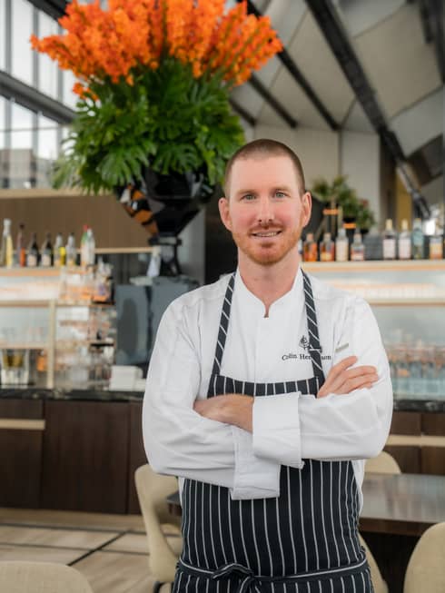 A chef wearing a white shirt and a striped apron stands in a brightly lit restaurant setting with tables and chairs, shelves, counters, and potted plants in the background.