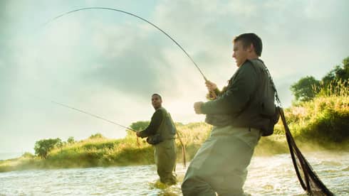 Two people fly fishing in a river, standing knee-deep in the water, surrounded by mist and greenery.