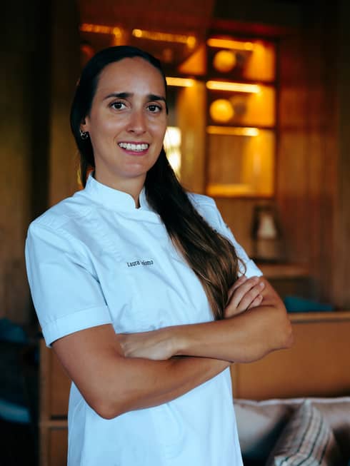 Chef with long brown hair and wearing a white, short-sleeve chef's shirt stands with her arms crossed smiling at the camera