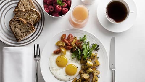 Breakfast plate with sunny-side-up eggs, cherry tomatoes, greens, and roasted potatoes, alongside toast, raspberries, grapefruit juice and coffee.
