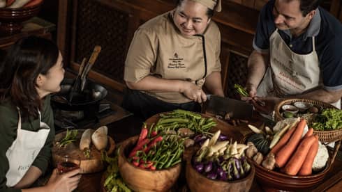 A chef teaching two participants in a cooking class, surrounded by fresh vegetables and ingredients in a rustic kitchen setting.