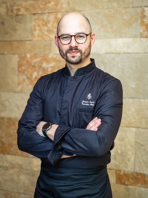 Person with shaved head, short beard and glasses stands with their arms crossed wearing an all black chef's uniform in front of a stone wall
