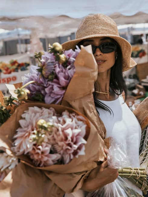 Person with long dark hair wearing a white sleeveless sundress, sunglasses and a floppy straw hat walks through a farmer's market holding three large bouquets of flowers