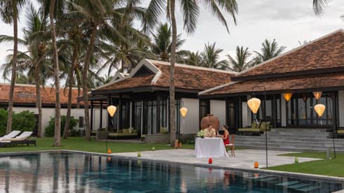 A couple enjoying a romantic dinner by a pool at a luxury villa, surrounded by palm trees and illuminated by hanging lanterns.