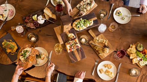 Aerial view of people dining around rustic wood table with platters of flatbreads, appetizers