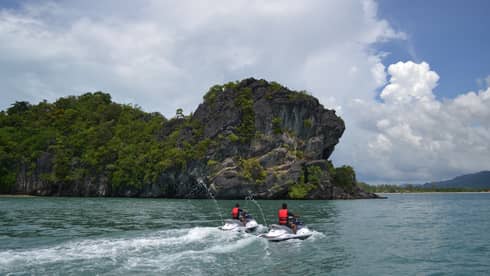 Two people wearing red life vests on jet skis ride on ocean under mountain