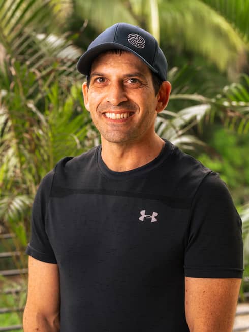 Person with short dark hair and wearing a black baseball cap and black t-shirt sits smiling in front of a green backdrop of palm trees