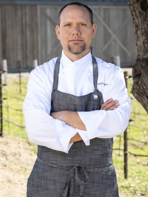 A chef standing outside near a tree and in front of a barn.
