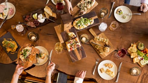 Aerial view of people dining around rustic wood table with platters of flatbreads, appetizers