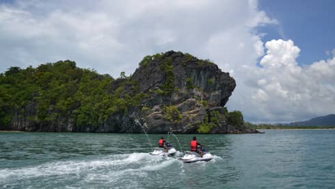 Two people wearing red life vests on jet skis ride on ocean under mountain